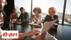 Im Hintergrund sind eine Gruppe von Mitarbeitern und Mitarbeiterinnen zu sehen. im Vordergrund das Logo von eon.
