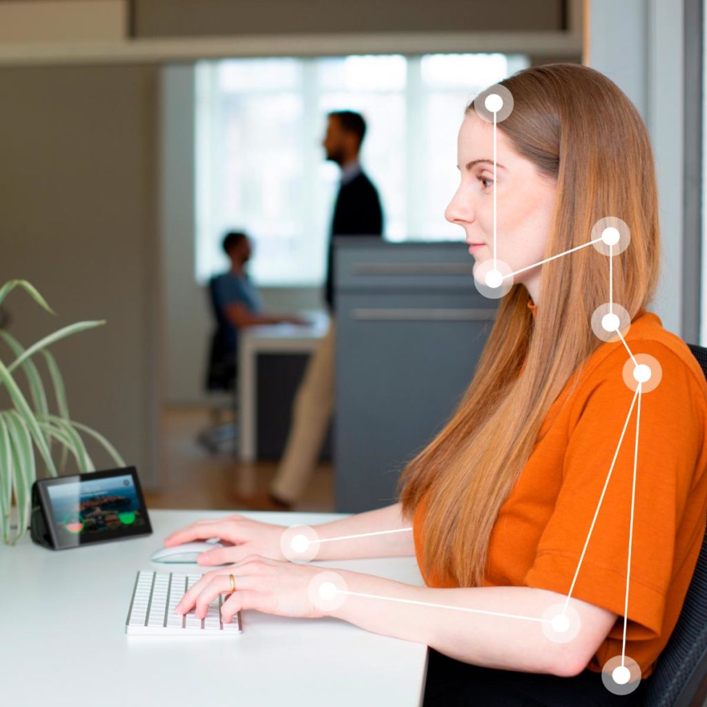 Eine junge Frau in einem orangenen Oberteil sitzt im Büro am Schreibtisch und arbeitet am Computer. Neben ihr steht eine Isa und misst die Ergonomie der jungen Frau. Durch Punkte und Striche auf dem Körper der Frau wird angezeigt, welchen Einfluss Isa auf die Körperhaltung hat.