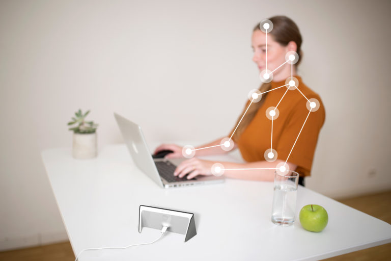 Mitarbeiterin sitzt am Schreibtisch und arbeitet am Laptop. Auf dem Schreibtisch steht ein Glas Wasser und ein Apfel. Daneben steht ein Isa Gesundheitscoach, welcher durch Sensorik die Körperhaltung der Frau analysiert. dies wird durch Punkte und Linien auf dem Körper der Frau angedeutet.