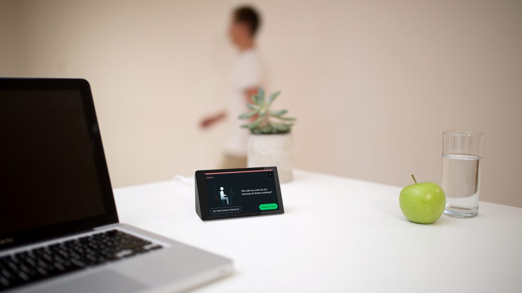 Das Bild zeigt Isa (die KI-Assistenz für gesunde Schreibtischarbeit) auf einem Schreibtisch neben einem Laptop