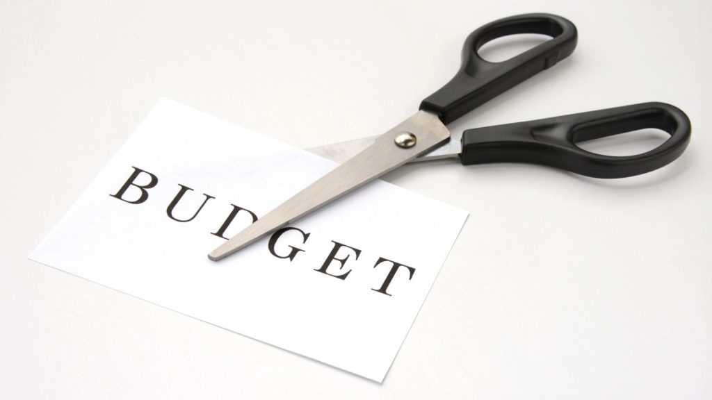 Zu sehen ist eine Schere, die ein Papier durchschneidet. Auf dem Papier steht Budget. 