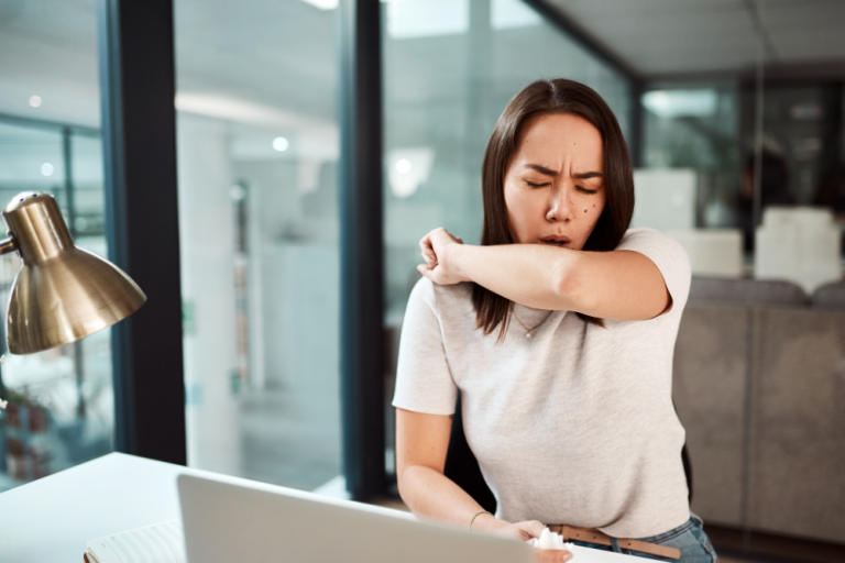 Eine Mitarbeiterin sitzt im Büro und hustet in ihre Ellenbeuge. Sie ist offensichtlich krank, aber dennoch im Büro und steckt unter Umständen Kollegen an.