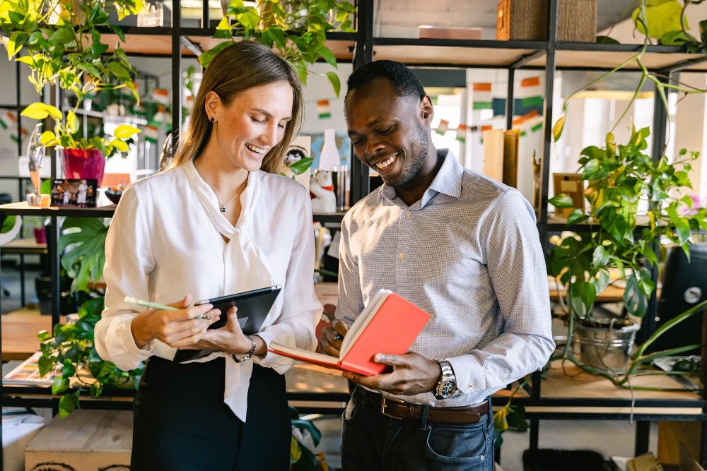 Zwei Mitarbeiter stehen zusammen im Büro und unterhalten sich über die Arbeit. Beide sehen glücklich aus, da die Präsentismuszahlen deutlich gesunken sind, nachdem Maßnahmen eingeführt wurden. 