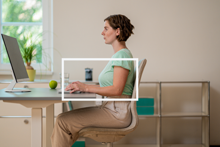 Zu sehen ist eine Mitarbeiterin an einem Schreibtisch. Der Fokus liegt auf der Armhaltung, die im 90 Grad Winkel ergonomisch korrekt ist. 