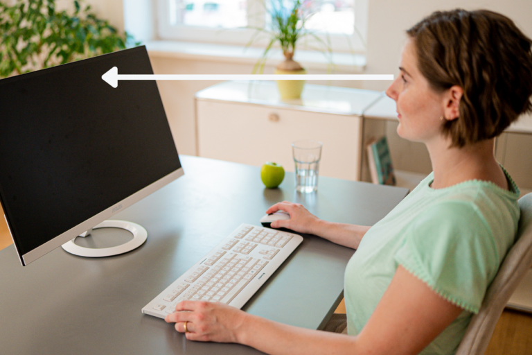 Zu sehen ist eine Mitarbeiterin am Schreibtisch, die auf einen externen Bildschirm schaut. Dieser Bildschirm soll eine Armlänge von den Augen entfernt sein und so stehen, dass der gerade Blickwinkel auf den oberen Teil des Bildschirms gerichtet ist.