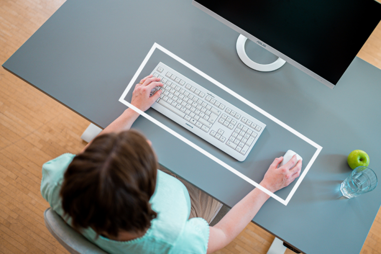 Zu sehen ist ein Schreibtisch mit Maus und Tastatur. Diese liegen flach und nicht weit von der Tischkante entfernt. 