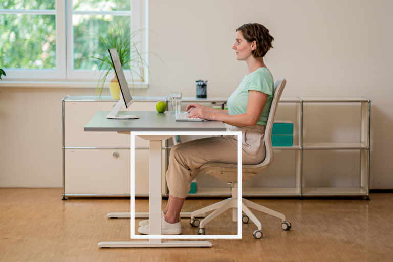 Zu sehen ist eine Mitarbeiterin an einem Schreibtisch. Im Fokus stehen die Beine. Die Füße stehen flach auf dem Boden und die Oberschenkel fallen leicht nach vorne ab. 